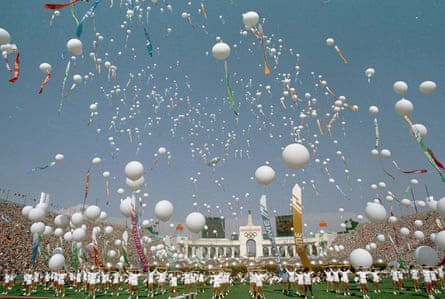 Participants stand on the field of a stadium and release white balloons into the blue sky