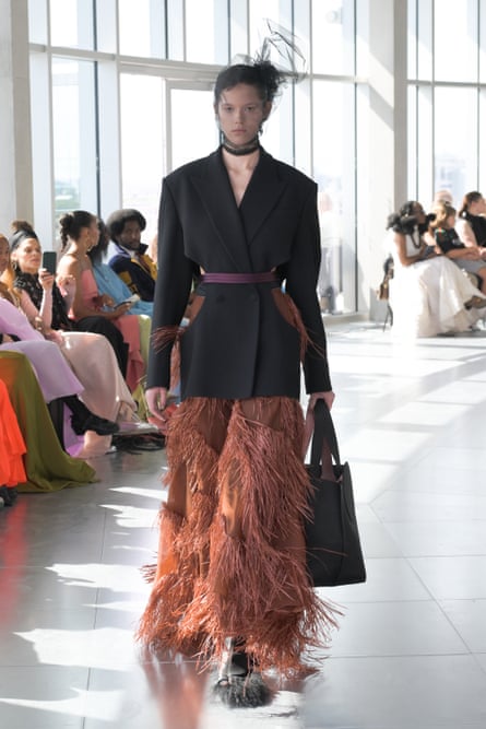 A model on the catwalk wearing a black blazer and fringed brown skirt.