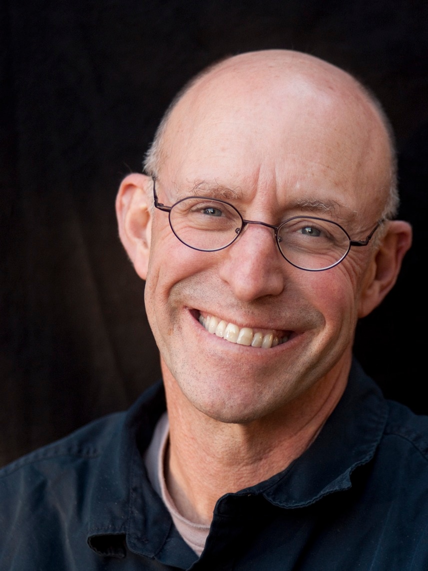 From the shoulders up, a bald man wearing dark blue shirt and thin-framed glasses smiles widely.