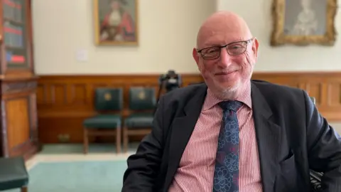 BBC Max Caller, lead commissioner at Birmingham City Council, sits inside an ornate room at the council's HQ. He looks at the camera and is wearing a pink shirt and dark grey suit jacket with a flower patterned tie.