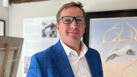 Grant Ford in glasses, blue jacket and white shirt in front of one of the paintings.