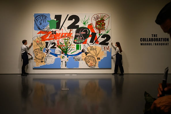 two art handlers stand holding up a massive canvas with the numbers 1/2 in black surrounded by abstract paintings