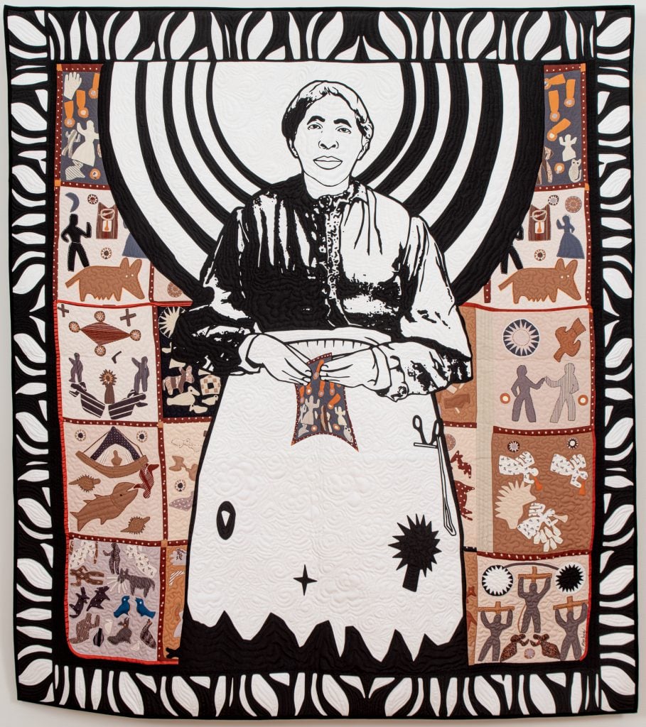 Carolyn Mazloomi, Mother of African QuiltingCarolyn Mazloomi Mother of African Quilting. A black and white art quilt with a brown background of quilting squares with African-inspired designs. The main figure is Harriet Powers, the first documented African American quilt artist. 