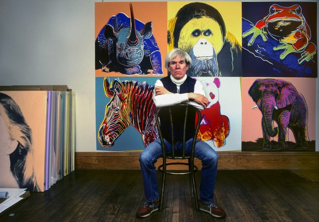 A white haired man sits in front of six pop art silkscreens of animals