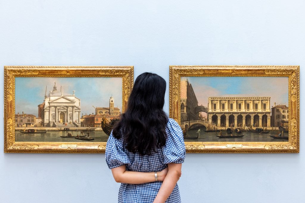 Two landscape paintings of Venice in gold frames hang on the wall in front of a dark haired woman