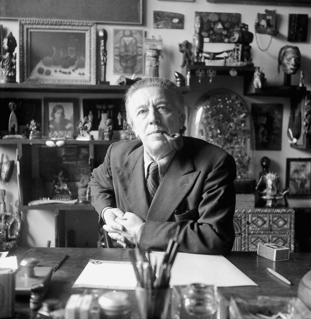 Black and white portrait of Andre Breton sitting at a desk in a cluttered office, who wrote one of the first Surrealist manifestos.