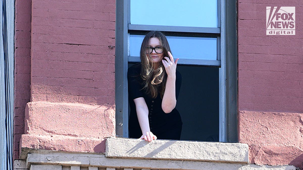 Anna Sorokin in a black top and glasses as she waves out a window of a brick building