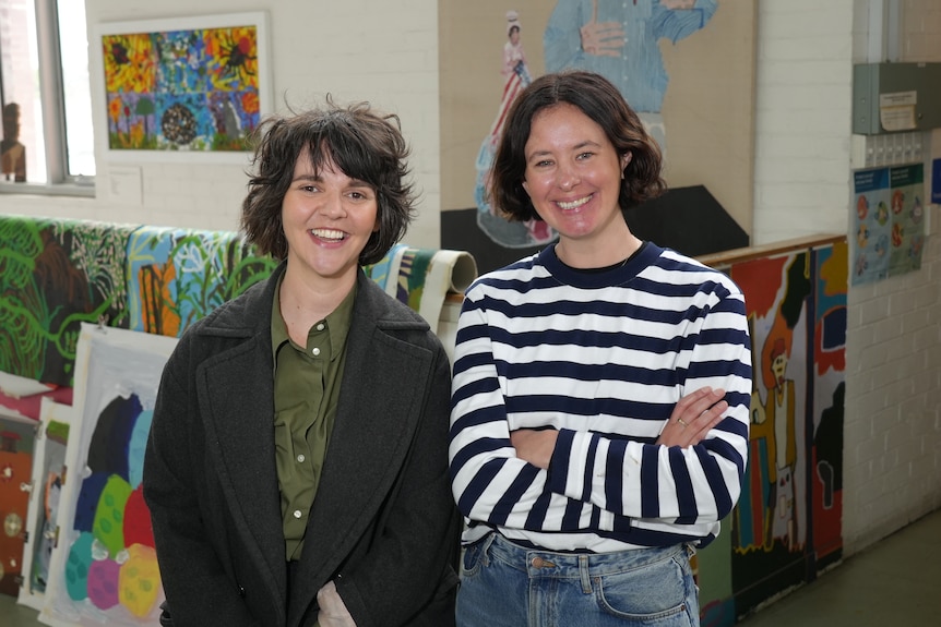 Two woman with short brown hair smile in front of artwork.