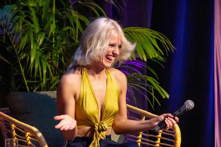 An image of Amy Taylor, who has blonde hair and wears a yellow top, smiling on a stage.