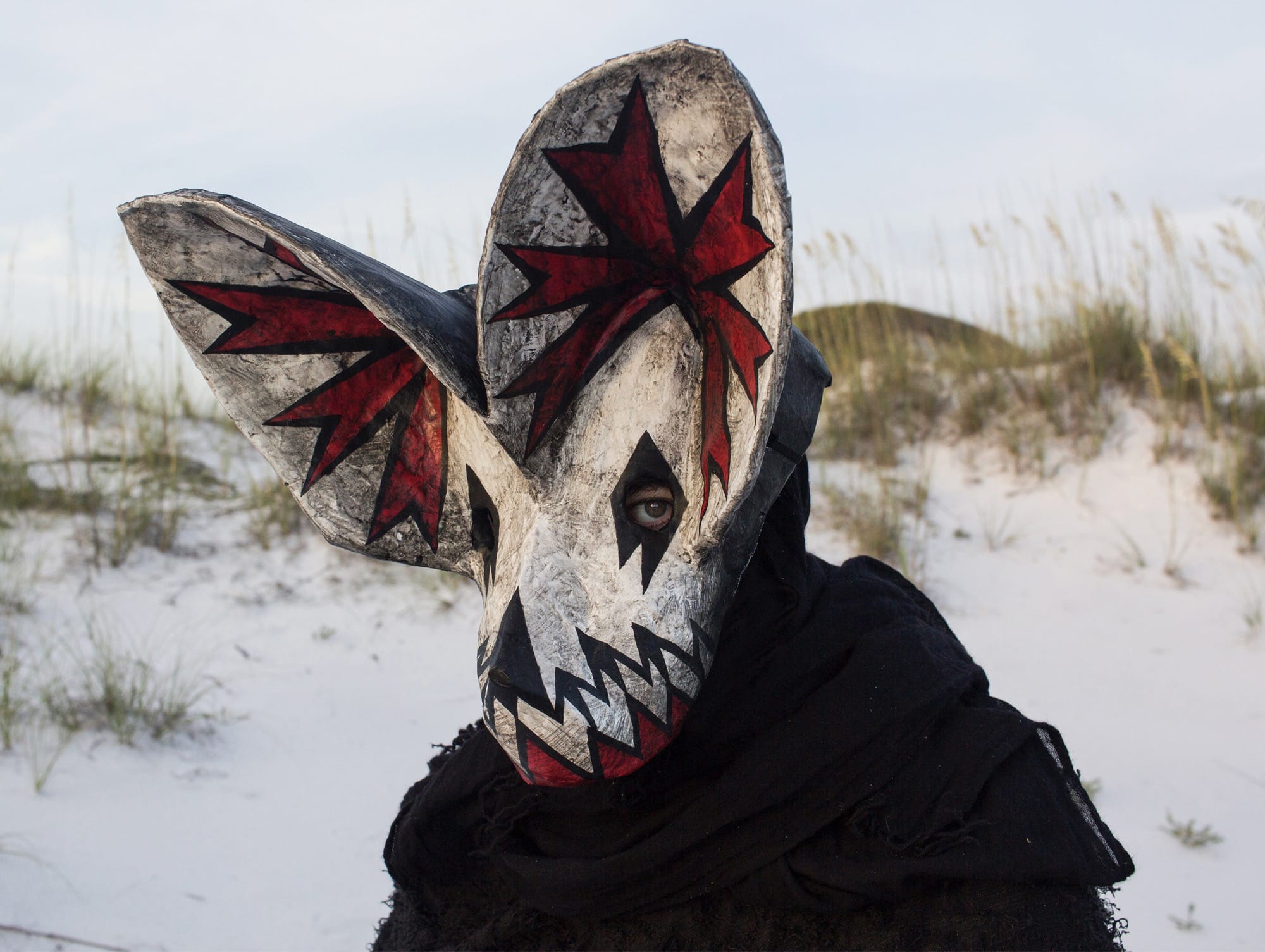 a portrait of a person wearing an ornate papier mache bat mask with red paint in the ears in a coastal setting