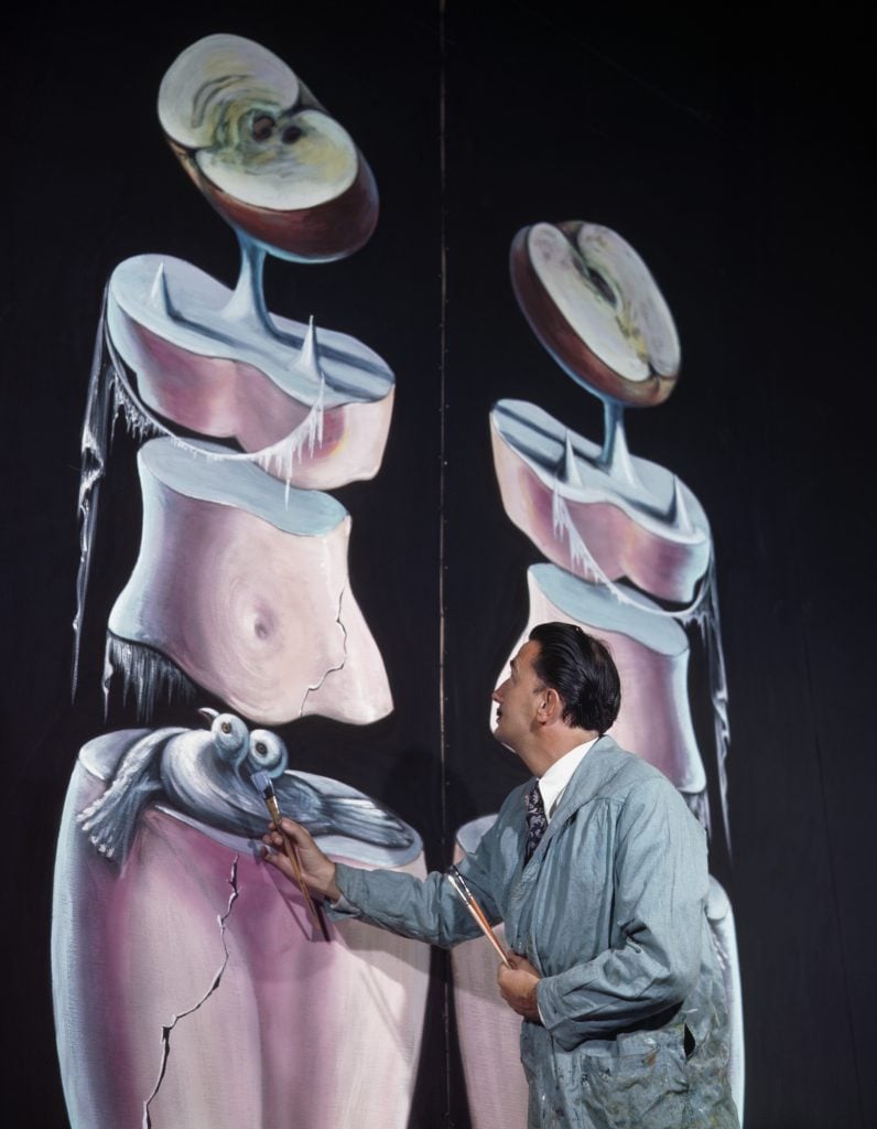 A color photo of Surrealist artist working on a large-scale Surrealist painting of two humanoid figures.