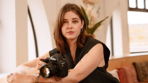 Phoebe Wingrove Artist Phoebe Wingrove sat with her camera. She is looking at the camera and wearing a short sleeved black top, with shoulder length brown hair. Behind her the room is decorated in neutral tones and there are large windows visible.