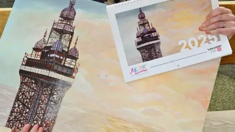 Lancashire County Council Kathryn Olley's winning painting features a close-up view of Blackpool Tower, set against a yellow and blue sky. Two seagulls flying above the sea reflects the same colours. In the top right of the picture we see a copy of the same painting on the front cover of a calendar for 2025.