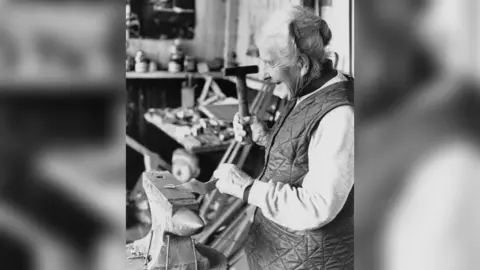 South West Heritage Trust An elderly woman holding a hammer, hammering a piece of metal on an anvil