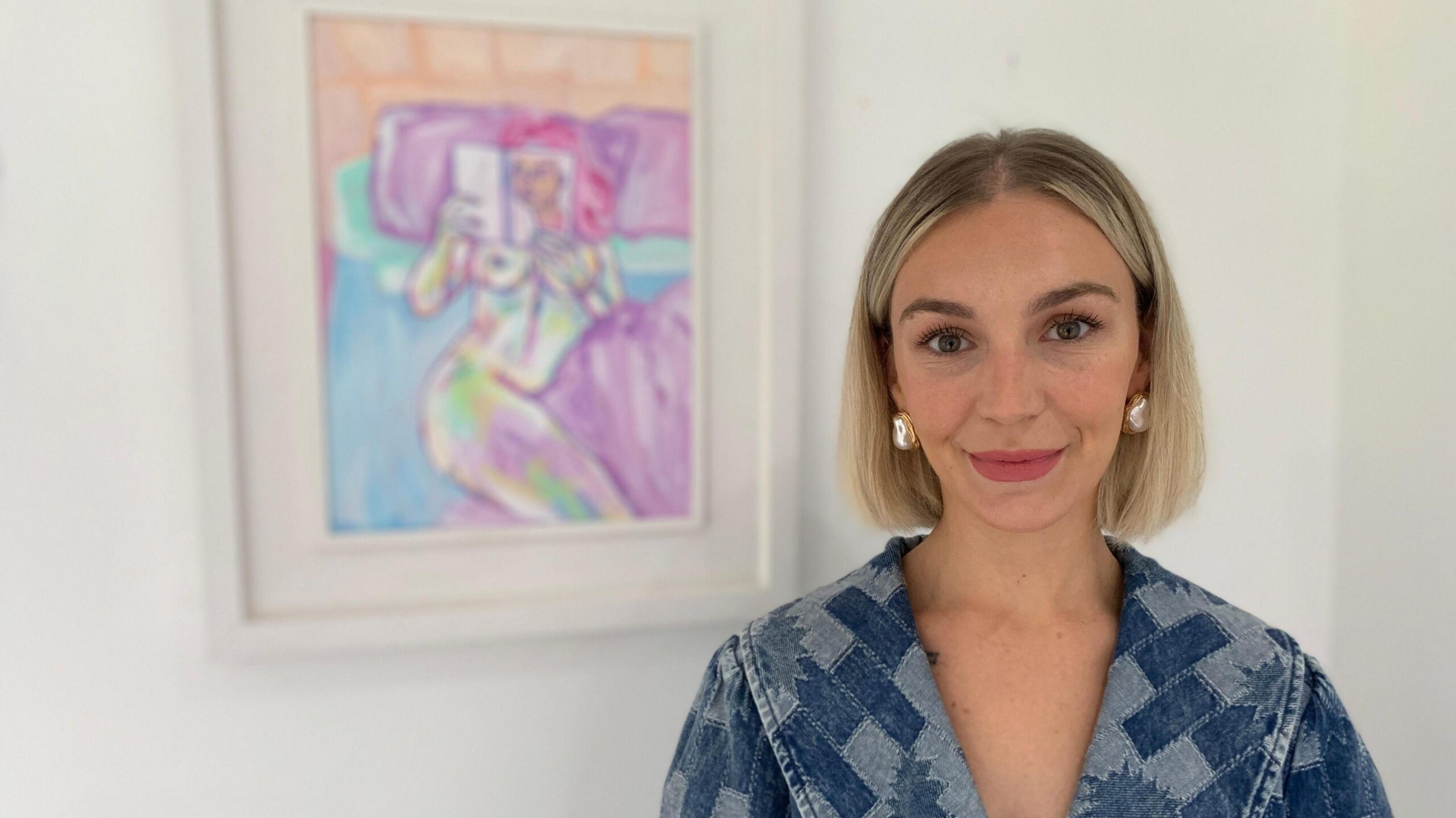 Clíodhna stands in front of her painting Laura. She has a blonde bob and is wearing a blue denim dress.