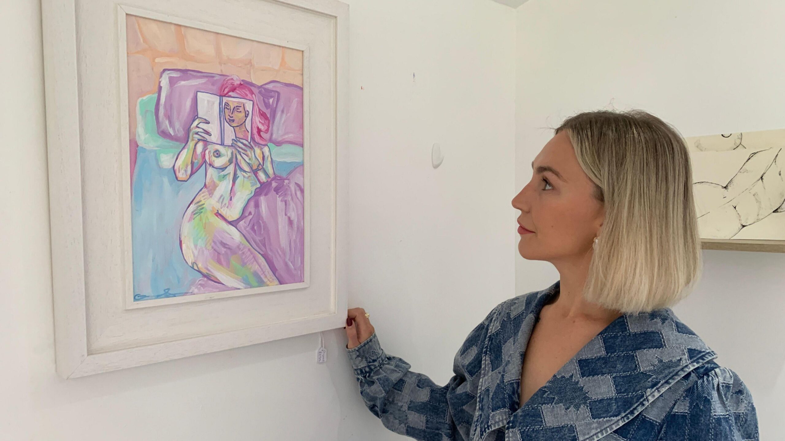 Clíodhna Doherty looks at her painting Laura in her art studio in Newry. The painting is a piece of abstract figurative art showing a naked woman reading a book while in bed. Clíodhna has a blonde bob and is wearing a blue denim dress.