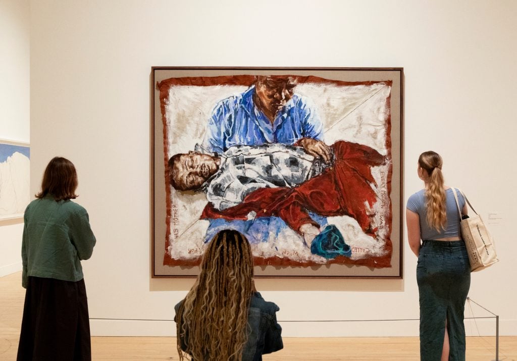 a photograph of a gallery space in which a large painting is being looked at by three onlookers, it is of a woman holding the body of a young boy in the classic biblical pose of Pieta