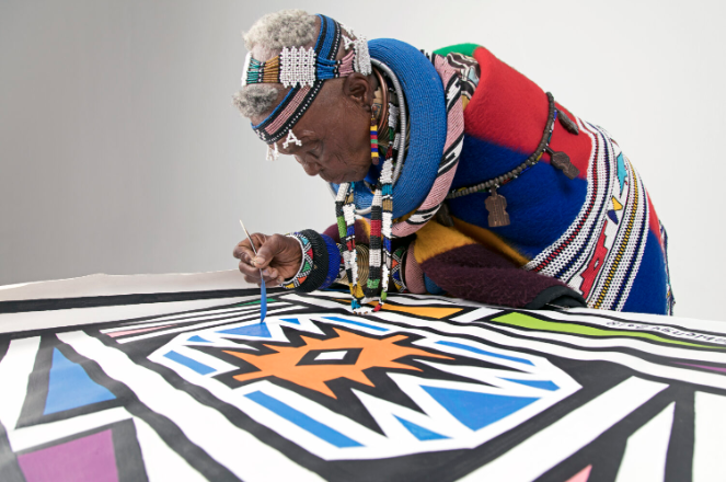 A woman of African descent dressed in colorful outfits and painting a colorful painting containing black borders