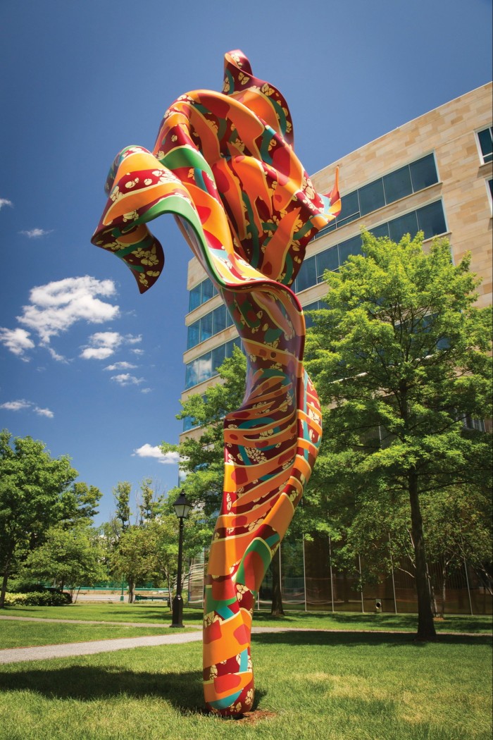 A large, vibrant outdoor sculpture, with swirling orange, red, green, and yellow patterns, stands on a grassy lawn