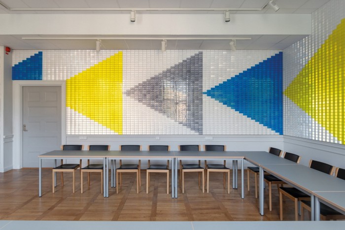 A bright, contemporary meeting room with a tiled wall design featuring large geometric triangles in yellow, blue, and gray, paired with neatly arranged gray tables and black chairs on a wooden floor