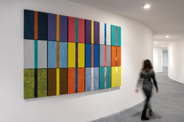 An abstract art piece with multicoloured vertical stripes on rectangular panels is mounted on a white wall in a modern hallway, with a blurred figure walking past