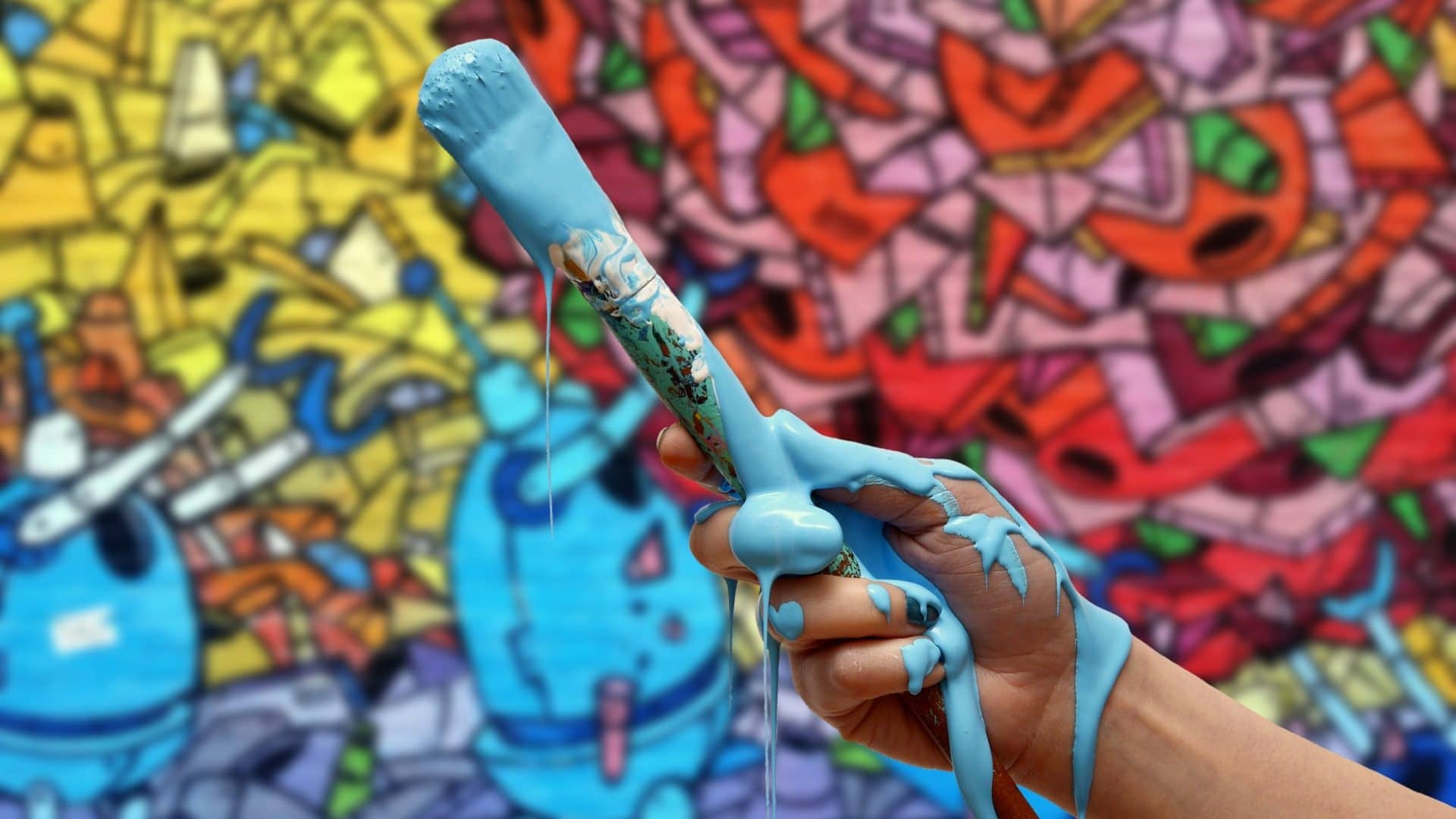 Image: Artistic person holding a paintbrush covered in paint in front of a wall with graffiti on it