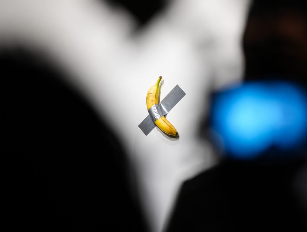  A single banana is duct-taped to a white wall as part of an art installation, with blurred dark silhouettes and a blue light in the foreground.