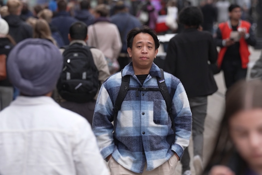 A man walking through a crowd, wearing a checked shirt and a backpack.