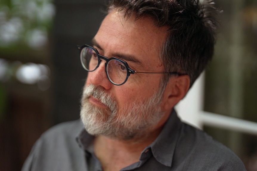 A close up of a man wearing glasses looking ahead as he sits on a balcony. His expression is contemplative.