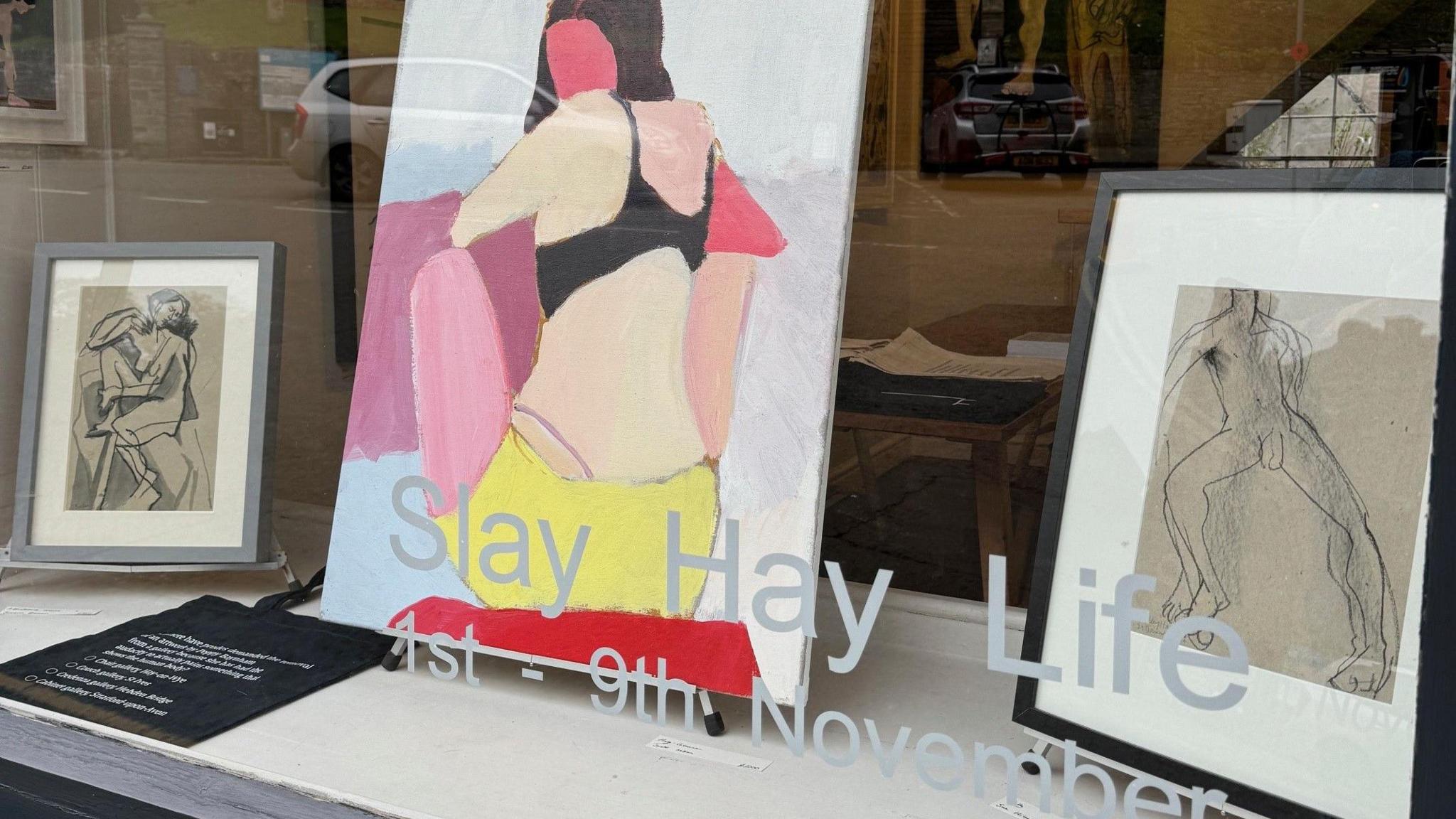 Picture of the gallery window display, one large canvas in centre of a woman's bare back, a framed line drawing of a naked man (right) and a framed image of a hunched over naked woman (left). 