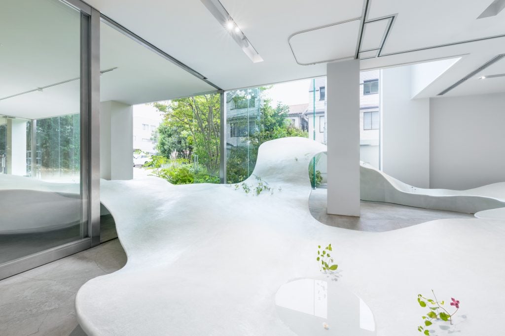 A white curvy landscape structure in a room, with green bushes at the back