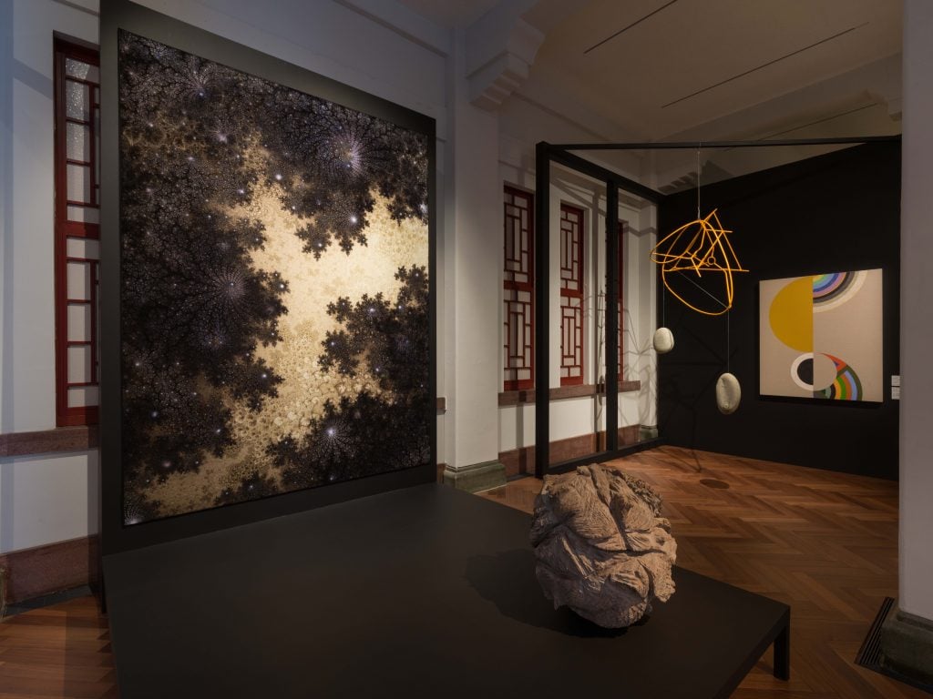 An abstract painting in black and beige hung on a podium, which has a rock placed in front of it, in an exhibition gallery of brown wooden floor. 