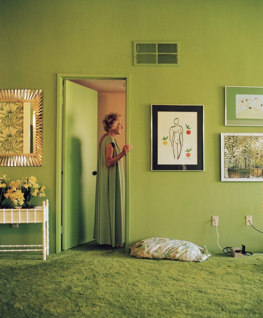 Larry Sultan, Mom in Green Nightgown (1992). An elderly women in a long green nightgown stands in the doorway of a bedroom painted the same color. There are artworks and a silver-framed mirror hanging on the wall, a pillow on the floor, and a table with a planting box full of daffodils. 