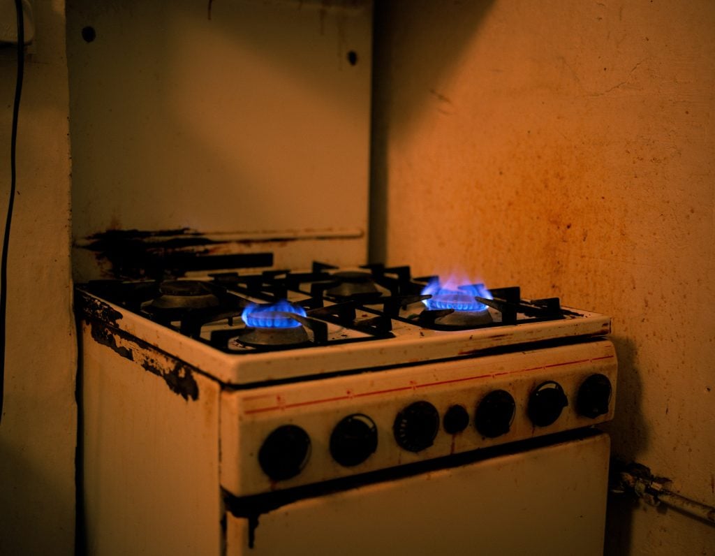 Paul Graham, Untitled (Flames), London (1998). A photo of a white gas stove in a dimly lit kitchen, the front two burner turned on and glowing blue.