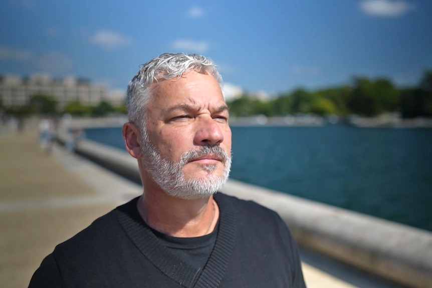 A man looks ahead as he walks on a footpath by a large body of water. He has a determined expression.