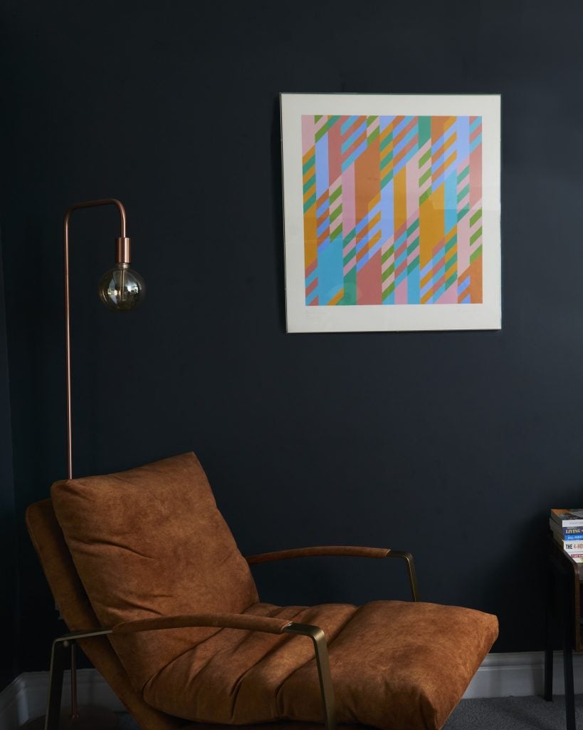 A pastel geometric abstract print by Bridget Riley installed on a dark navy wall behind a brown suede chair, on offer from Lougher Contemporary