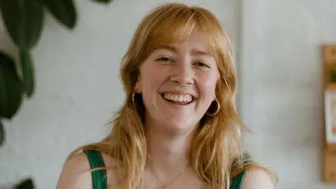 Rachael Reid Ana Jaks has strawberry blonde hair and is wearing a green strappy top and gold earrings. She smiles at the camera.