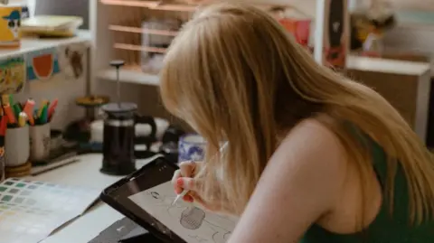 Rachael Reid Ana sits at a desk which has stationary placed on it. She is working on an illustration on an iPad.