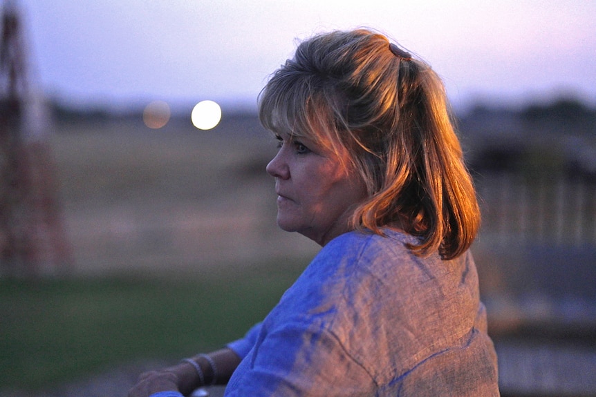 A woman stands leaning on a railing at dusk looking ahead, in the distance are fields. Her expression is sad.