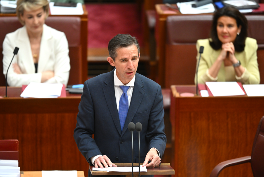 A man in a navy suit with a blue tie stands before a microphone