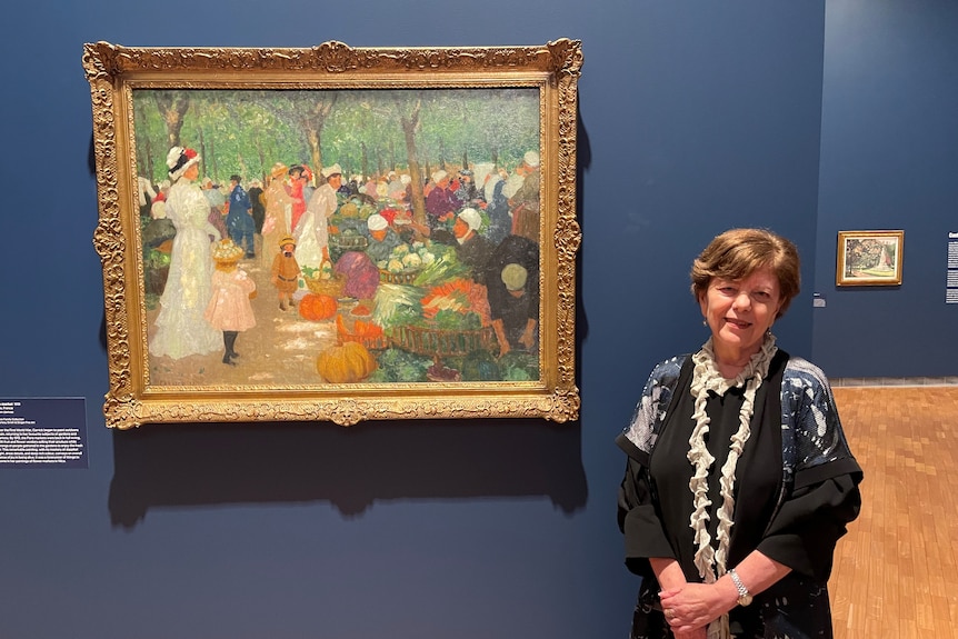 A woman with short hair stands next to a post-impressionist painting of people in Edwardian clothing at an outdoor market.