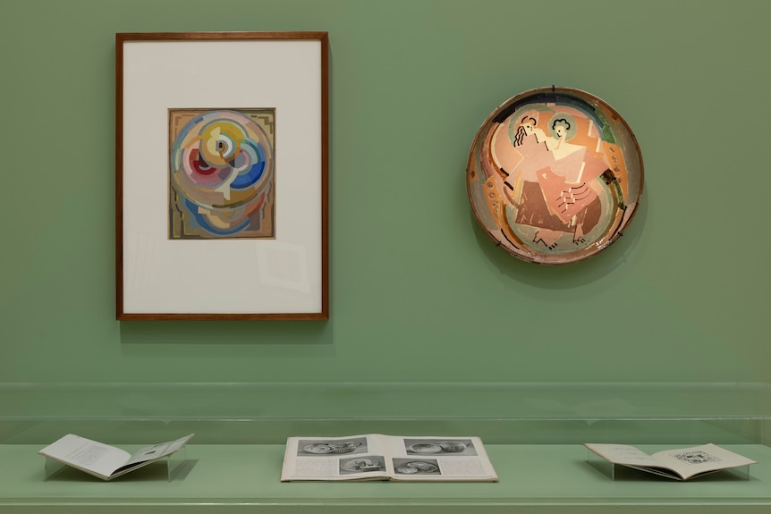 A cubism painting and circular ceramic piece hanging on a gallery wall.