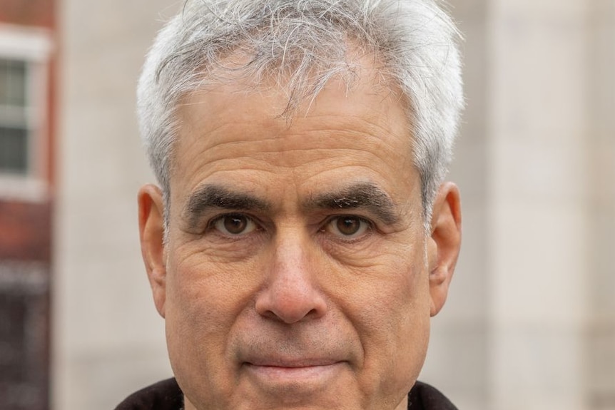 A man with light grey hair smiles in front of an building, outdoors.