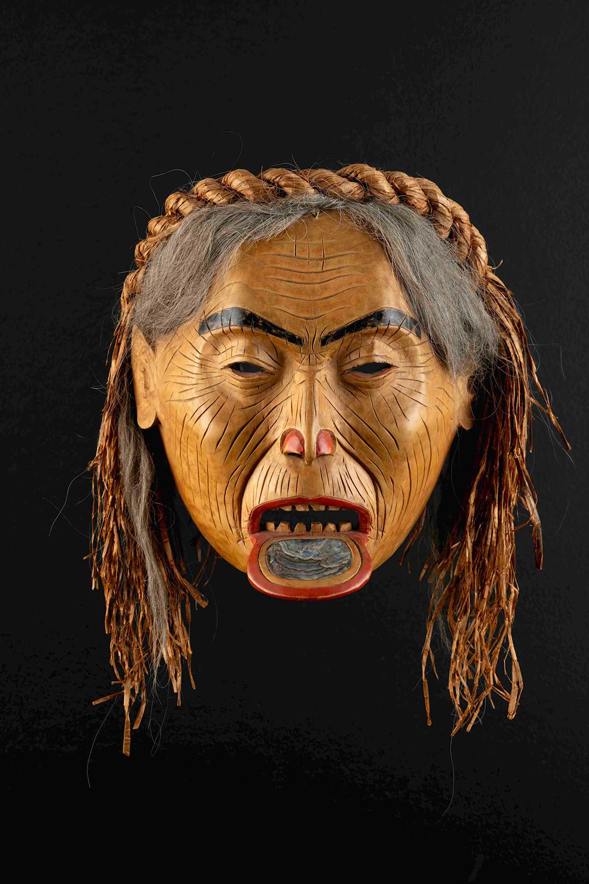 A wood mask depicts an older woman’s face with long grey hair. She is wearing a braided cedar bark headpiece and has a labret piercing adorned with abalone shell.