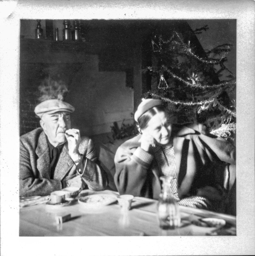 A black-and-white photograph showing an older man smoking a cigarette and a woman sitting beside him, both at a table. A Christmas tree stands in the background, and the woman rests her head on her hand, appearing pensive, while the man gazes off to the side with smoke rising above him.