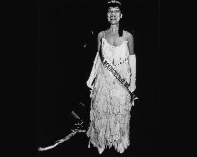 A Black woman wearing a dress made out of gloves with a sash reading 'BOURGEOISE' running across it. She shouts before a black background.