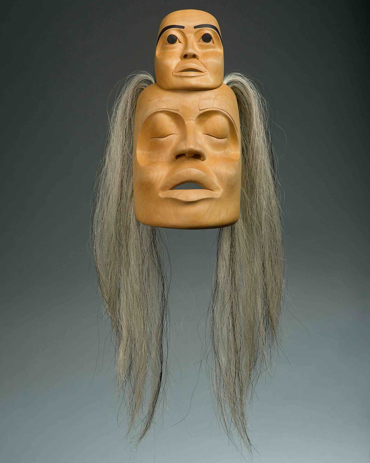 A carving of a woman’s face with long grey hair; a smaller face sits atop her forehead.