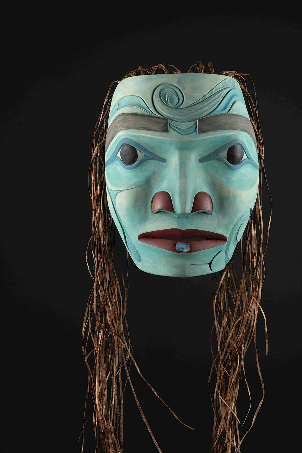 A wood mask features a woman’s face in turquoise. Her face is surrounded by long strands of cedar bark that suggest hair.