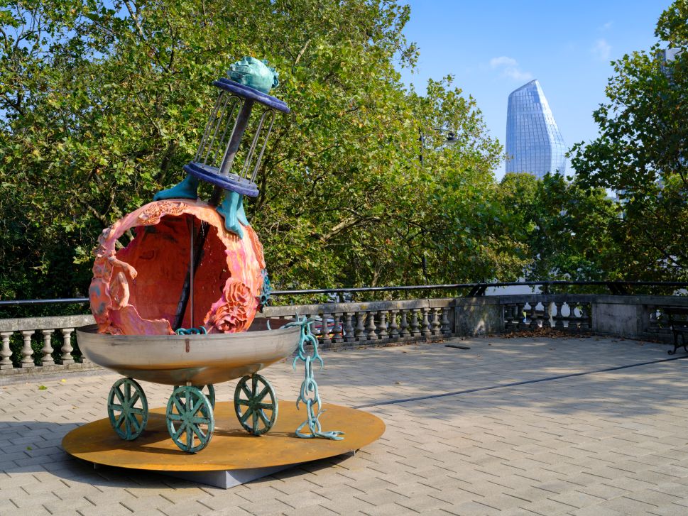 A photo of a whimsical sculpture featuring a partially open orange sphere mounted on wheels, with a blue figure holding a cage-like structure on top. The artwork is displayed on a paved terrace, surrounded by trees, with a tall modern glass building visible in the background.