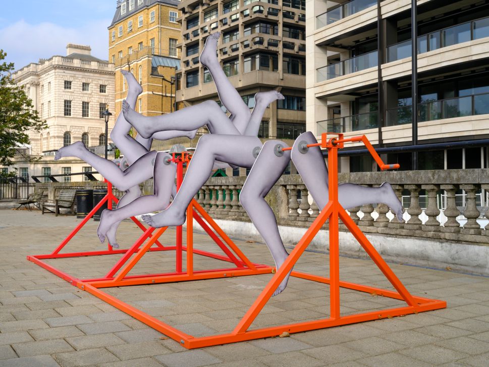 A photo of an abstract sculpture featuring multiple disjointed mannequin legs attached to an orange metal frame with cranks. The artwork is positioned on a paved surface, with classical and modern buildings visible in the background.